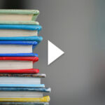 Photo: A stack of hardcover books of various colors. There is a white play button overlayed on top of the image